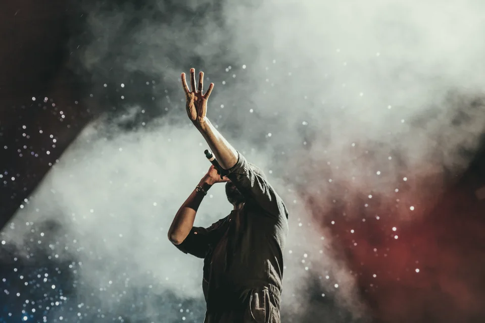 A vocalist holding a microphone raises one hand in the stage lights.